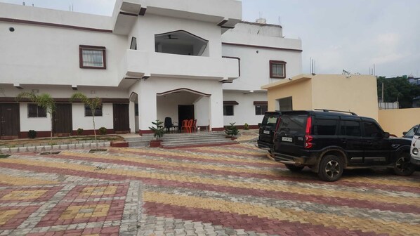 Un edificio de varios pisos con una entrada central, un patio empedrado y dos SUVs estacionados.