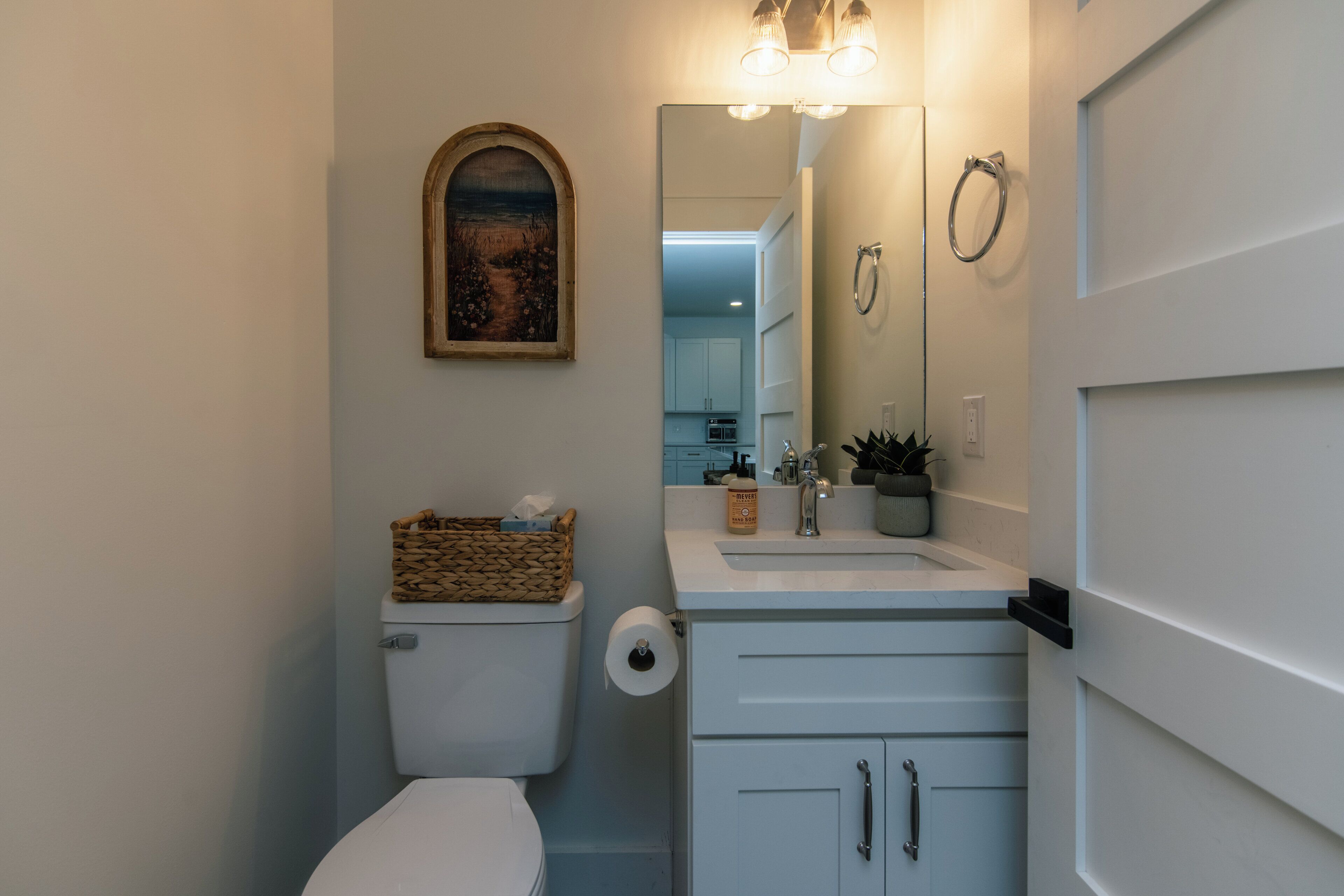 A bathroom with a toilet, a sink with a mirror, a basket on the toilet tank, and a towel ring on the wall.