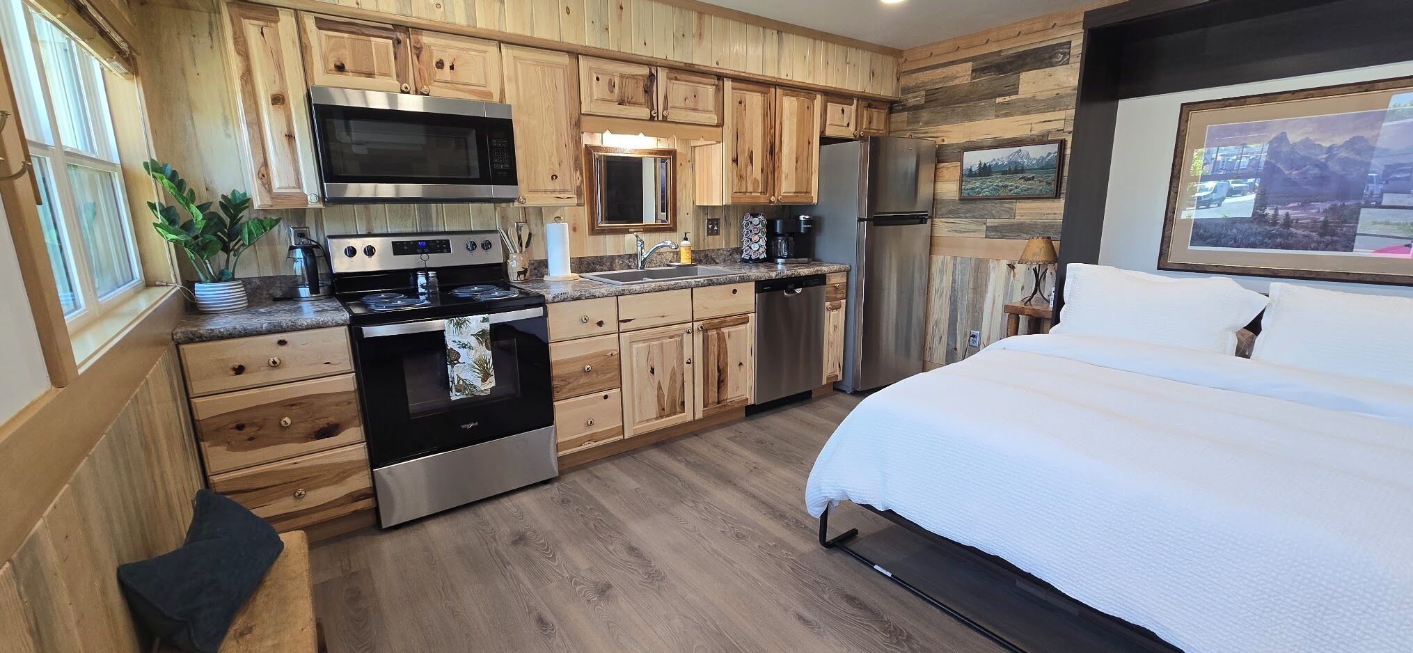 A kitchen with wooden cabinets, a microwave, stove, refrigerator, and a bed with white bedding.