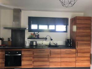 Una cocina moderna con armarios de madera, una campana extractora de acero inoxidable, horno empotrado, cafetera y una ventana con persianas.