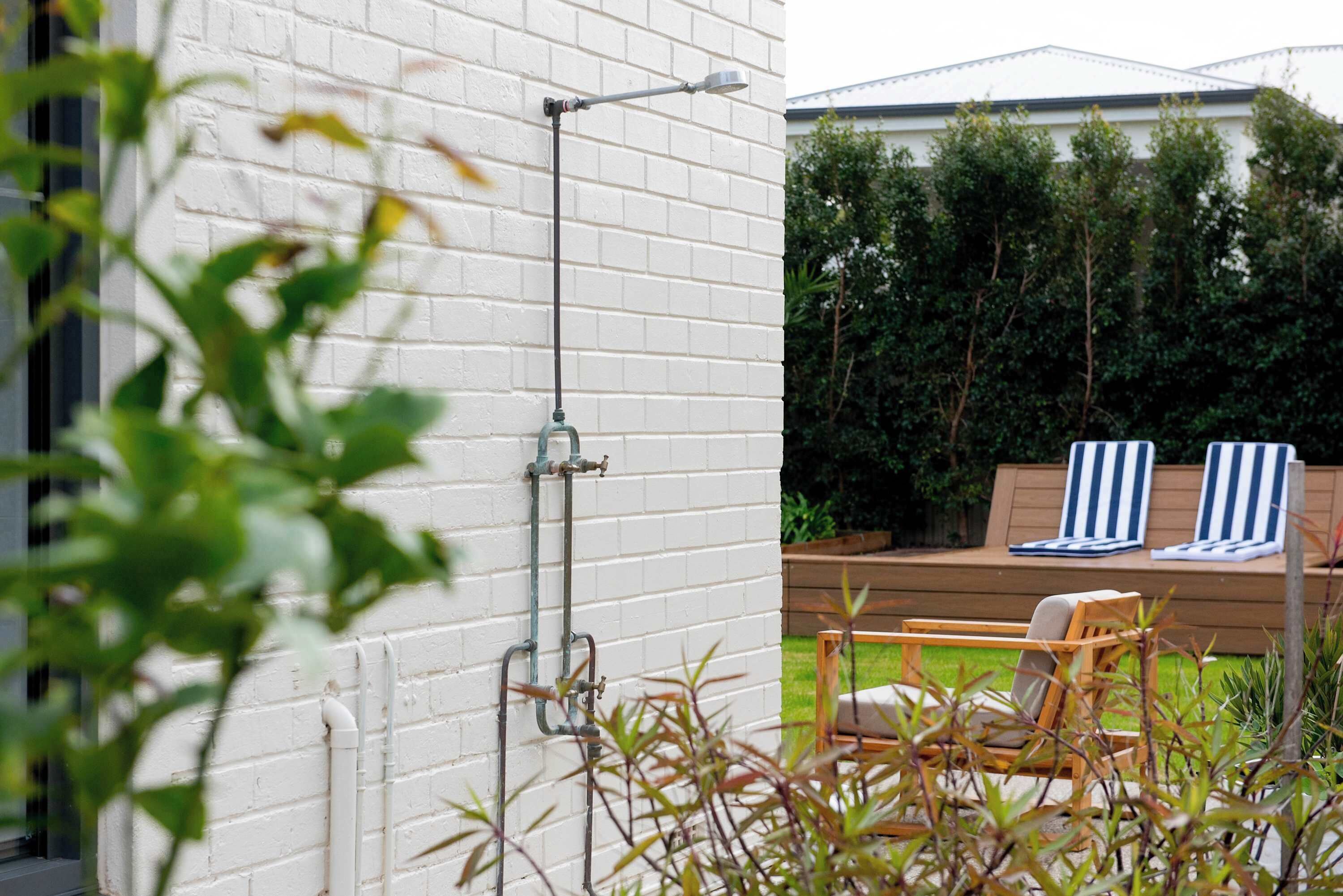 A white brick wall with outdoor plumbing, a wooden deck with two striped lounge chairs, and a green hedge in the background.