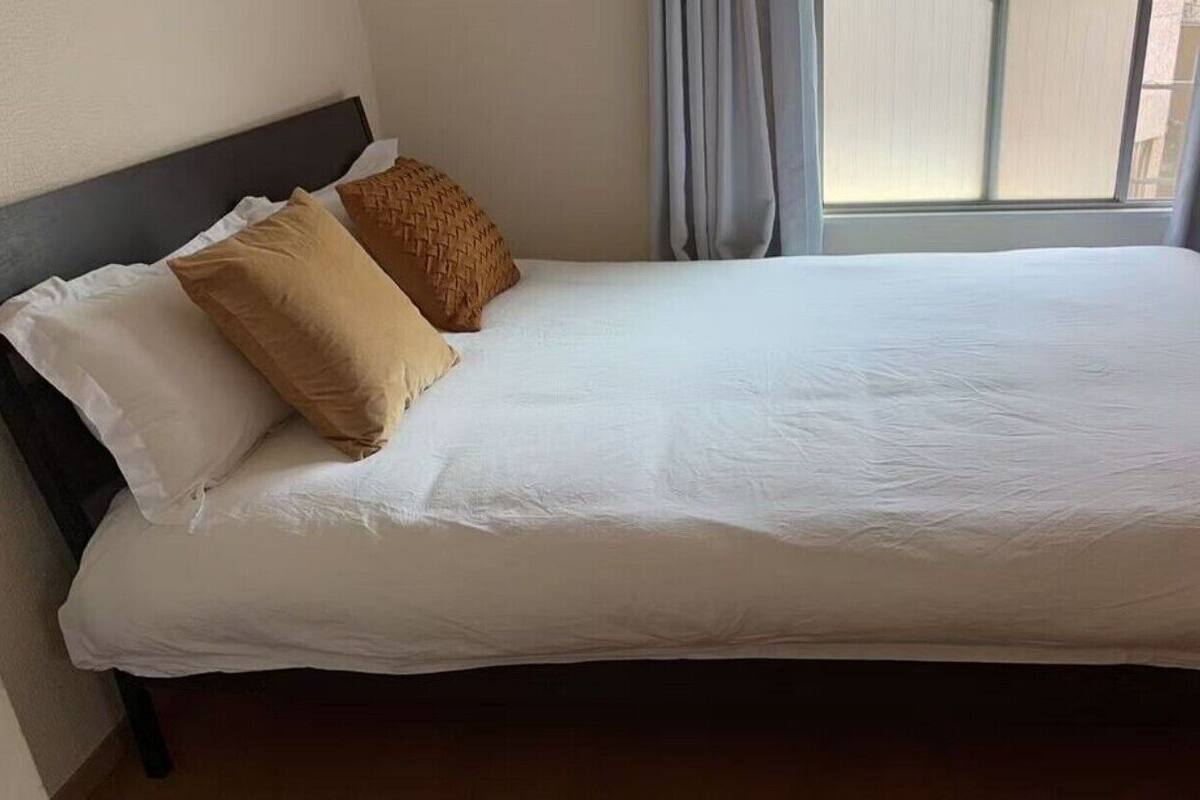 Una cama bien hecha con ropa blanca y dos almohadas, un cabecero oscuro y una ventana con cortinas.