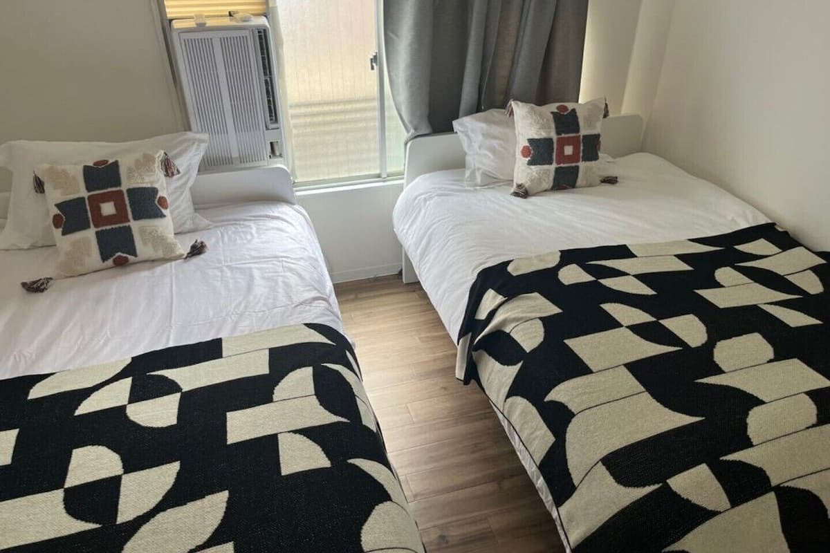 Una habitación con dos camas, cada una con una colcha estampada, un radiador, una ventana con cortinas y un suelo de madera.