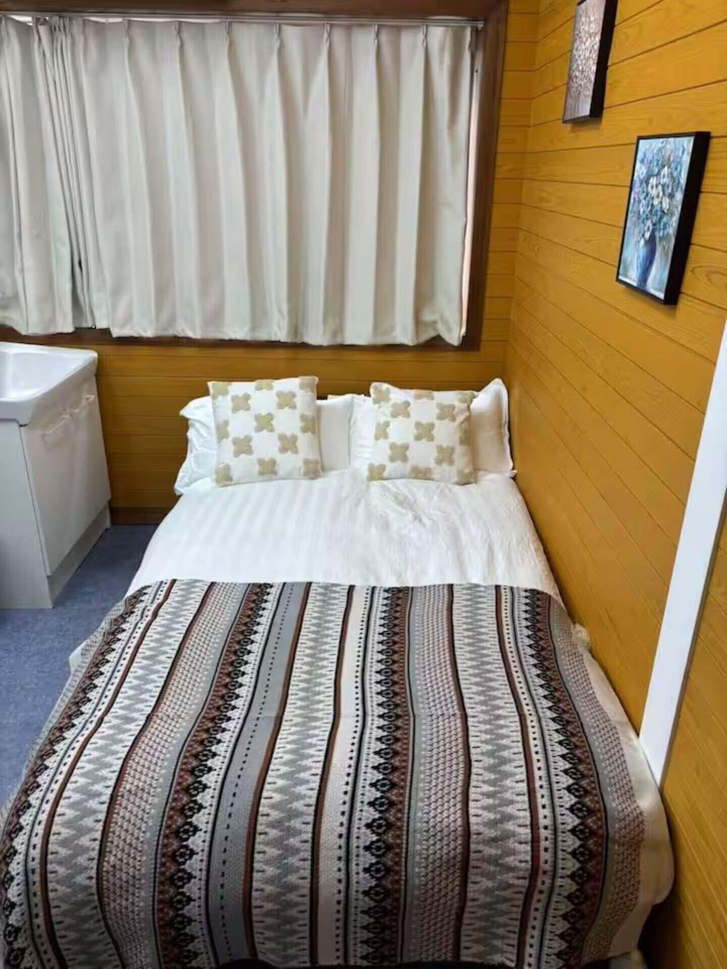 A small, cozy room with a bed, a wooden wall, a window with curtains, and a sink.