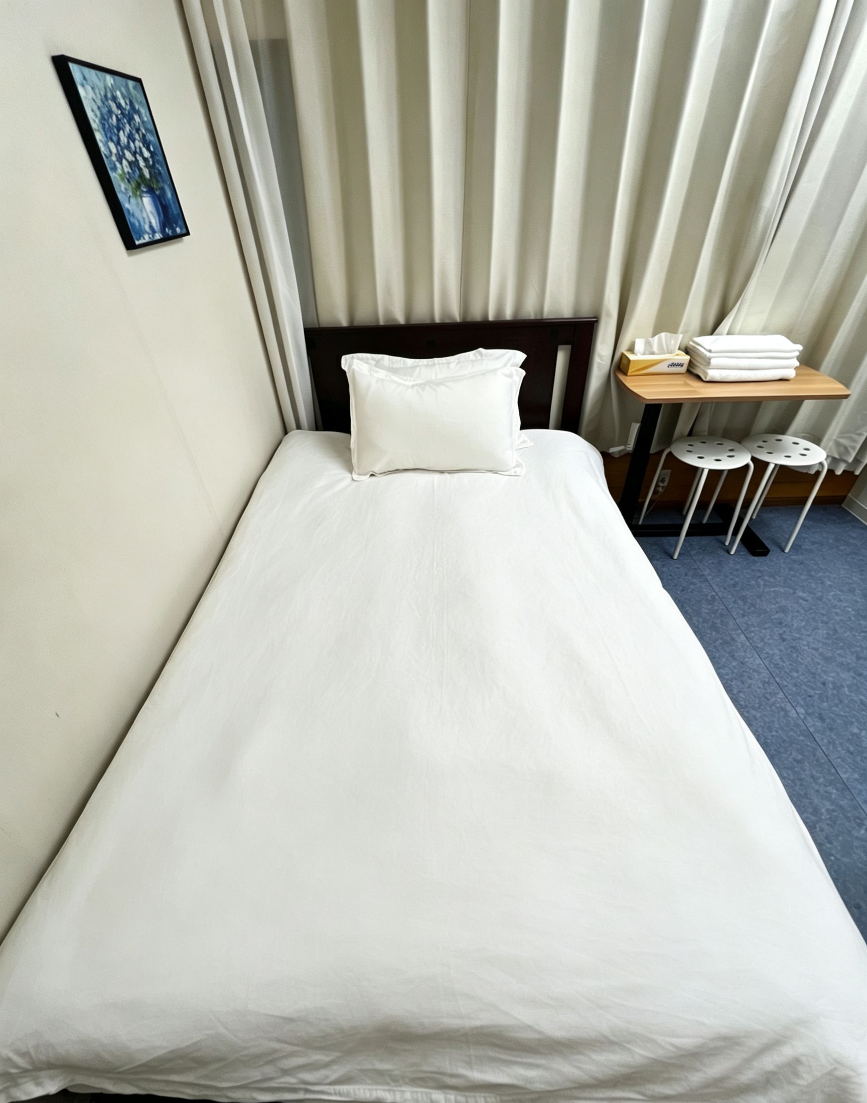 Uma cama de solteiro com roupa de cama branca, cabeceira de madeira, uma pequena mesa com toalhas e um banquinho.
