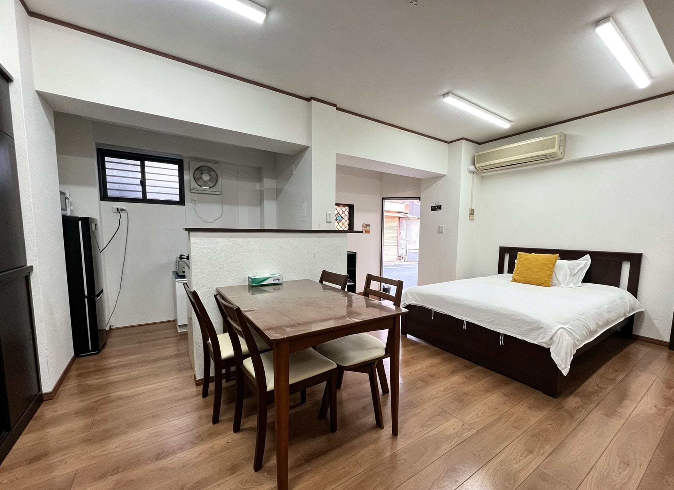 Una habitación compacta con una mesa de comedor de madera, sillas, una cama con colchón blanco y almohada amarilla, y un equipo de aire acondicionado en la pared.