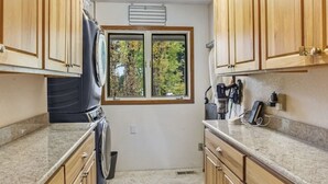 A compact kitchen with wooden cabinets, a built-in washer and dryer, a window with a view of trees, and granite countertops. - 2-bedroom house in tranquil Grapeview perfect for relaxing getaways (Grapeview)