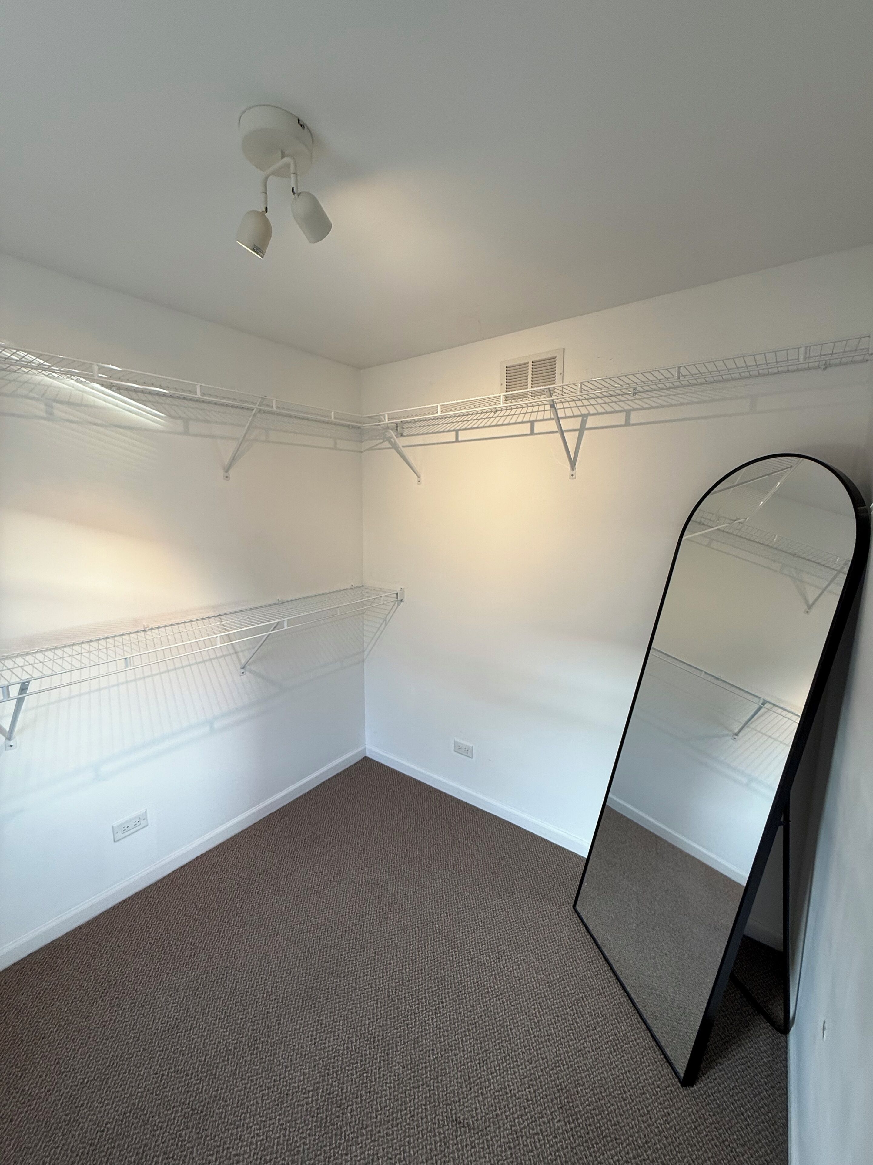 Ein Zimmer mit Deckenleuchte, Drahtregal, Spiegel und Teppichboden.