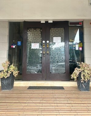 Entrada de hotel com porta de madeira escura, painéis de vidro e dois vasos de plantas de cada lado.