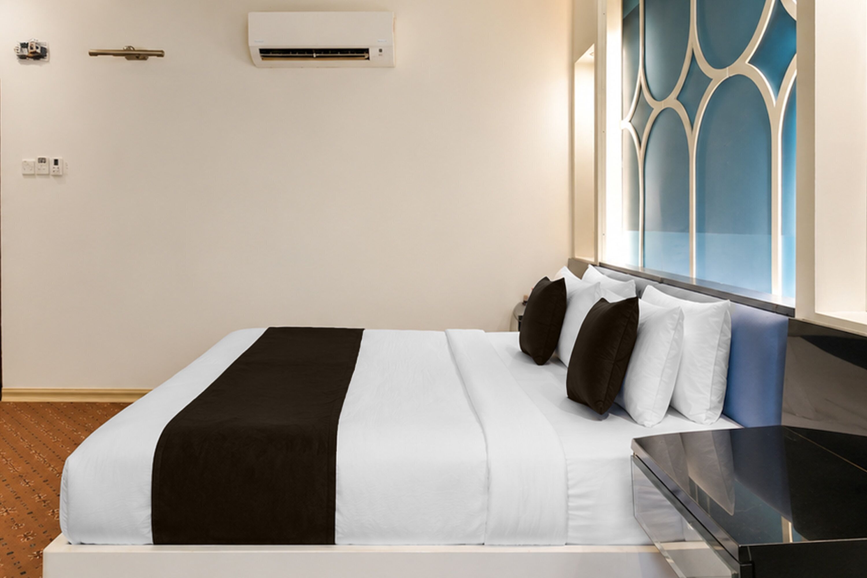 A hotel room with a bed, a nightstand, an air conditioning unit, and a window with a decorative view.