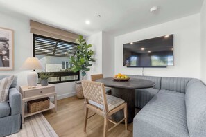 Ein modernes Wohnzimmer mit einem grauen Sofa, einem Holzstuhl, einem Esstisch mit Obst, einem an der Wand montierten Fernseher und einem Fenster mit Jalousien.