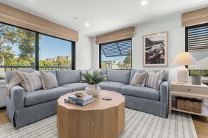 Ein modernes Wohnzimmer mit einem grauen Ecksofa, einem runden Holztisch und großen Fenstern mit Blick ins Freie.