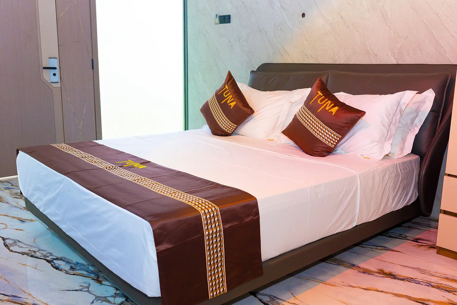 A hotel room with a bed, white bedding, brown decorative pillows, and a marble-patterned floor.