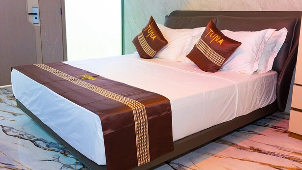 Habitación de hotel con cama, ropa de cama blanca, almohadones decorativos marrones y piso con estampado de mármol.