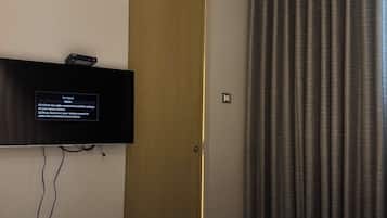 A hotel room with a flat-screen TV mounted on the wall, a door with a golden frame, and a large window with dark curtains.