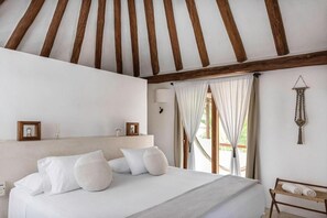 Une chambre à coucher avec un lit, des poutres en bois, une table de chevet, une lampe fixée au mur et une fenêtre avec des rideaux.