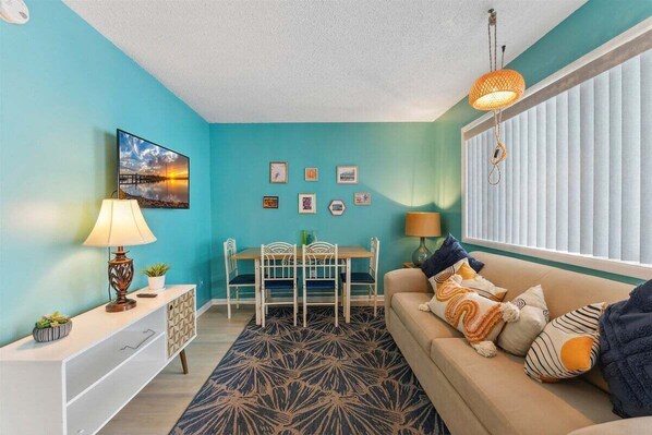 A living room with teal walls, a sofa with patterned cushions, a dining table with chairs, and a hanging light fixture.