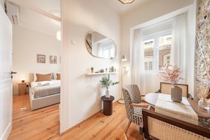 Ein Zimmer mit einem Bett, einem Sessel, einem Tisch, einem Spiegel und einem Vorhangfenster.