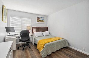 Un dormitorio con cama, escritorio con computadora, silla y ventana con persianas.