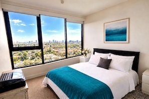 A hotel room with a large window, a bed with white bedding, a black headboard, a small bedside table with a vase of flowers, and a framed picture on the wall. - Deluxe 2-Bed with Stunning Melbourne Views & Central South Yarra Location (South Yarra)