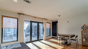 Una habitació amb un llit, una taula de dinar amb cadires, una estanteria i grans finestres amb cortines.