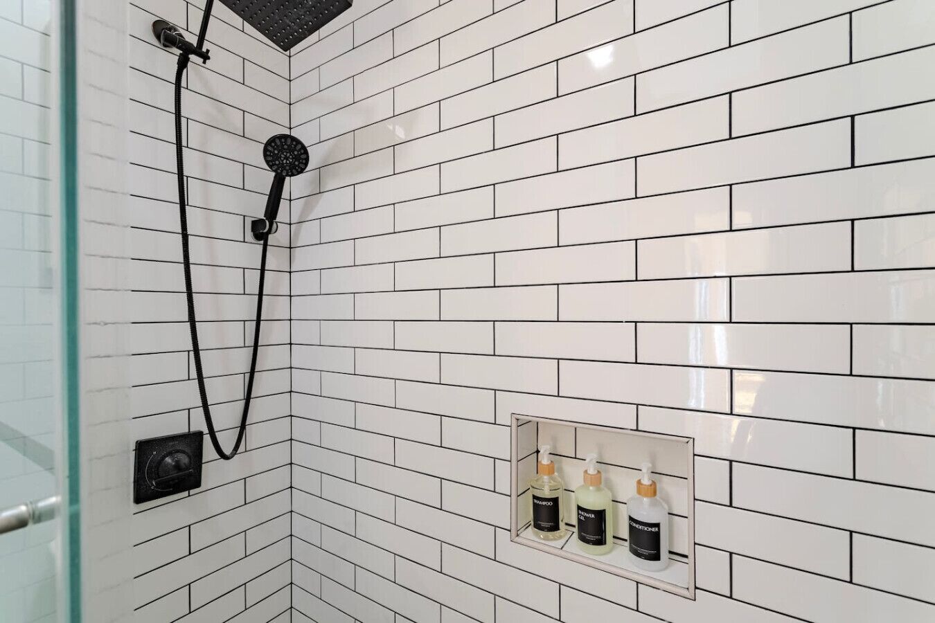Área de duche com paredes revestidas a azulejos brancos, chuveiro manual e prateleira com três frascos de produtos de higiene.