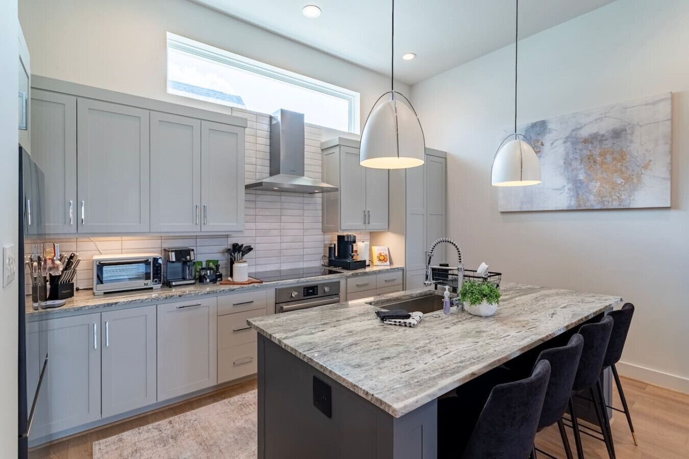 Une cuisine moderne avec des meubles gris, un îlot central, des luminaires suspendus et une grande fenêtre.