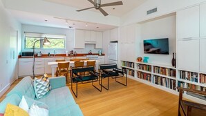 A modern kitchen with white cabinets, a dining area with a table and chairs, a sofa, and a bookshelf with books and decorative items. - Home for 4 near i10 / Memorial / Bayou Trails (Houston)