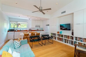 A modern kitchen with white cabinets, a dining area with a table and chairs, a sofa, and a bookshelf with books and decorative items. - Home for 4 near i10 / Memorial / Bayou Trails (Houston)
