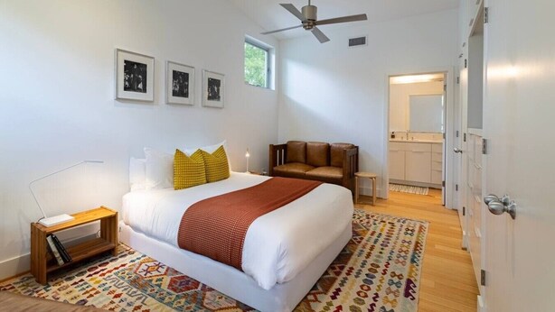 A modern bedroom with a bed, a ceiling fan, a small seating area, a patterned rug, and a bathroom visible through an open door. - Home for 4 near i10 / Memorial / Bayou Trails (Houston)