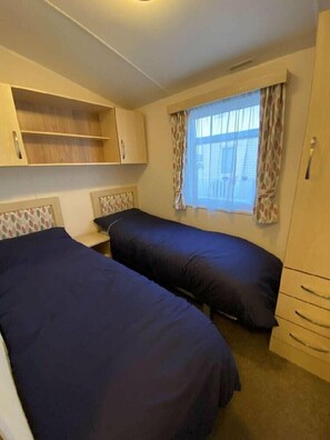 A room with two beds, a window with curtains, a wardrobe, and a shelf unit. - Lyons Robin Hood - Holiday Accommodation 17224 (Rhyl)