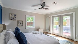 Un dormitorio con cama, ventilador de techo, ventana, espejo y puerta corredera de vidrio que da a un área exterior.