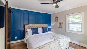 Un dormitorio con un cabecero capitonado, ventilador de techo, ventana con persianas y cama con ropa de cama blanca y almohadas azules.