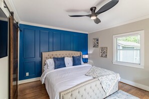 Ein Schlafzimmer mit einem gepolsterten Kopfteil, einem Deckenventilator, einem Fenster mit Jalousien und einem Bett mit weißer Bettwäsche und blauen Kissen.