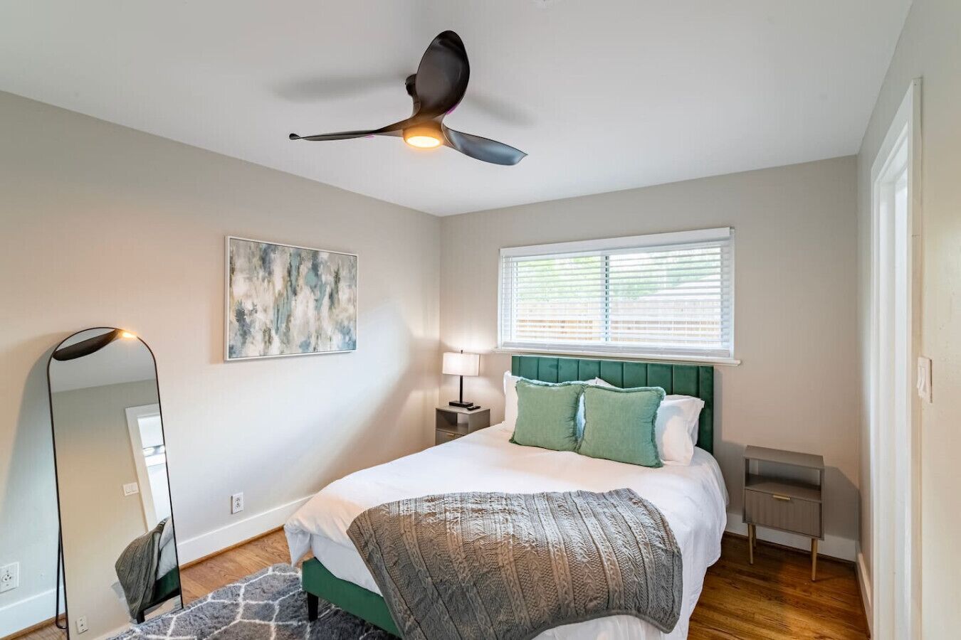 A bedroom with a bed, a ceiling fan, a bedside table with a lamp, a wall-mounted mirror, and a window with blinds.