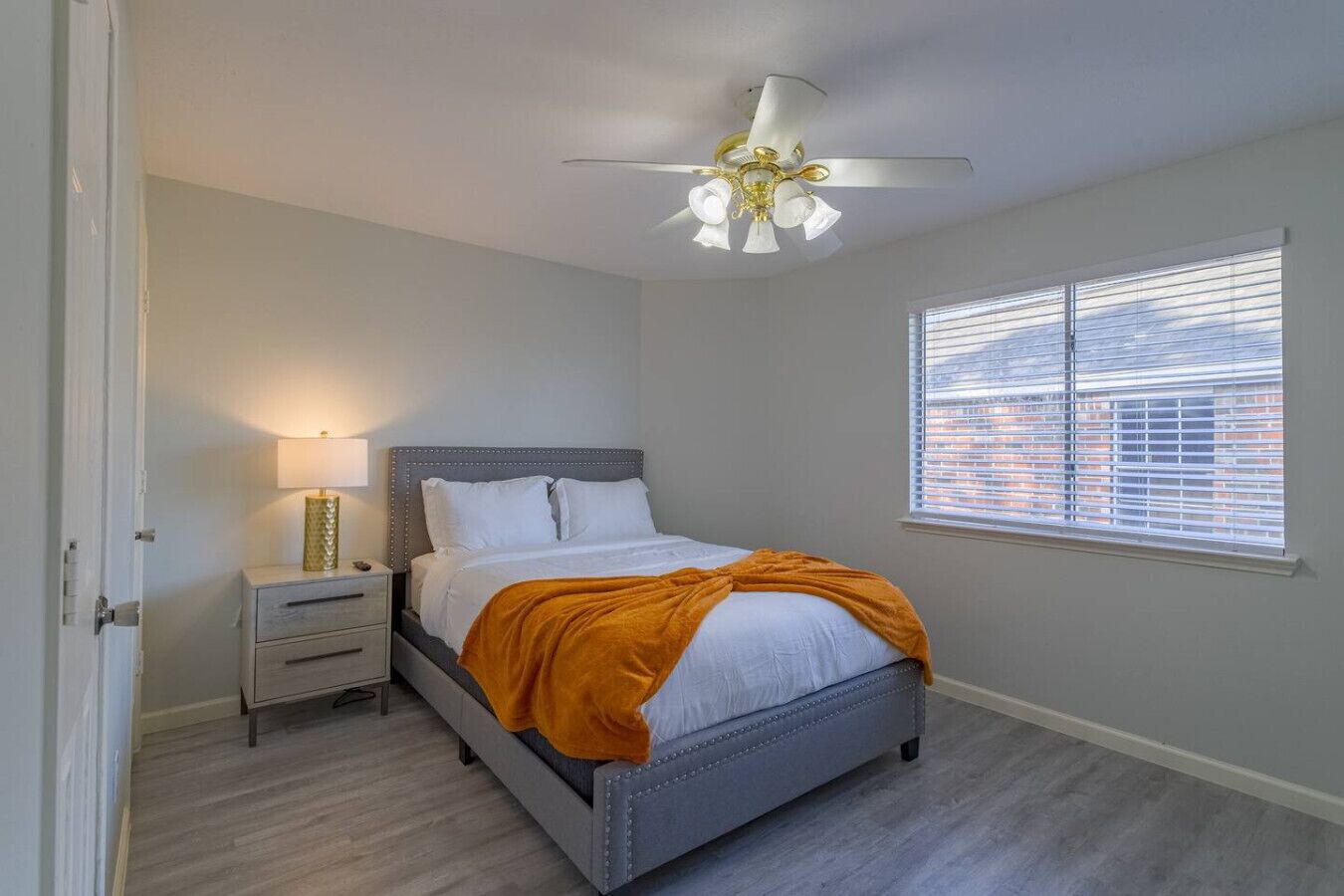 Een slaapkamer met een bed, nachtkastje, lamp, plafondventilator en raam met jaloezieën.