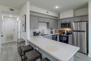 A modern kitchen with grey cabinets, a stainless steel refrigerator, a microwave, and a large island with stools. - Cozy NRG apt for 5 near NRG stadium (Houston)
