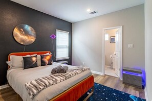 Quarto com cama, mesa de cabeceira, relógio na parede, janela com persianas e porta para um banheiro.