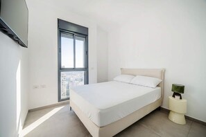 Um quarto com uma cama, janela, TV fixada na parede e mesa de cabeceira com abajur.