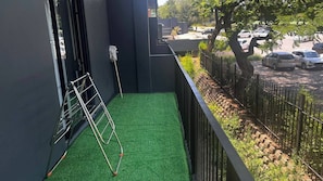 Ein Balkon mit Kunstgrashalm, einer Metallleiter und Blick auf geparkte Autos und Grünflächen.