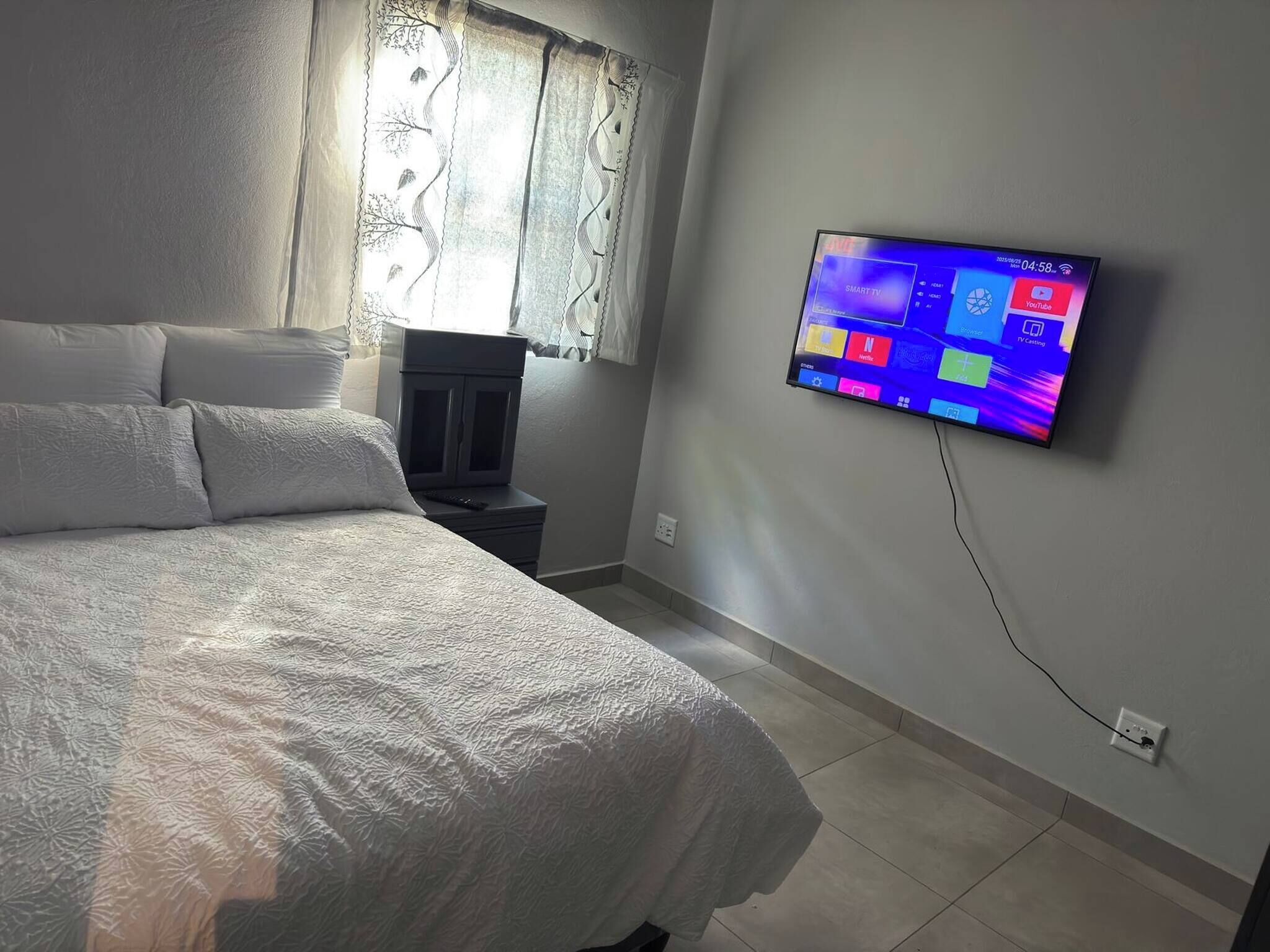 Ein Schlafzimmer mit Bett, Wandfernseher, Fenster mit Vorhängen und Nachttisch mit Fernbedienung.