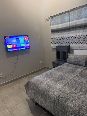 Ein Schlafzimmer mit einem Bett, einem an der Wand montierten Fernseher, einem Nachttisch und einem Fenster mit gemusterten Vorhängen.