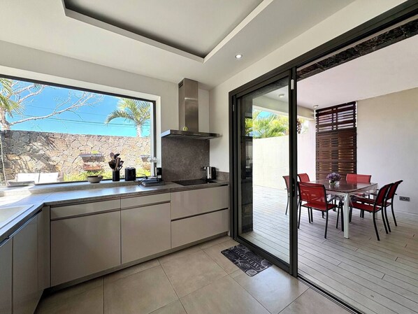 A modern kitchen with a large window, a dining area with red chairs, and a stone wall outside. - Modern Villa Nestled in its Tropical Garden (Grand-Baie)