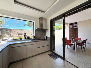 A modern kitchen with a large window, a dining area with red chairs, and a stone wall outside. - Modern Villa Nestled in its Tropical Garden (Grand-Baie)