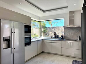 A modern kitchen with stainless steel appliances, a stone backsplash, and a window with a view of greenery. - Modern Villa Nestled in its Tropical Garden (Grand-Baie)
