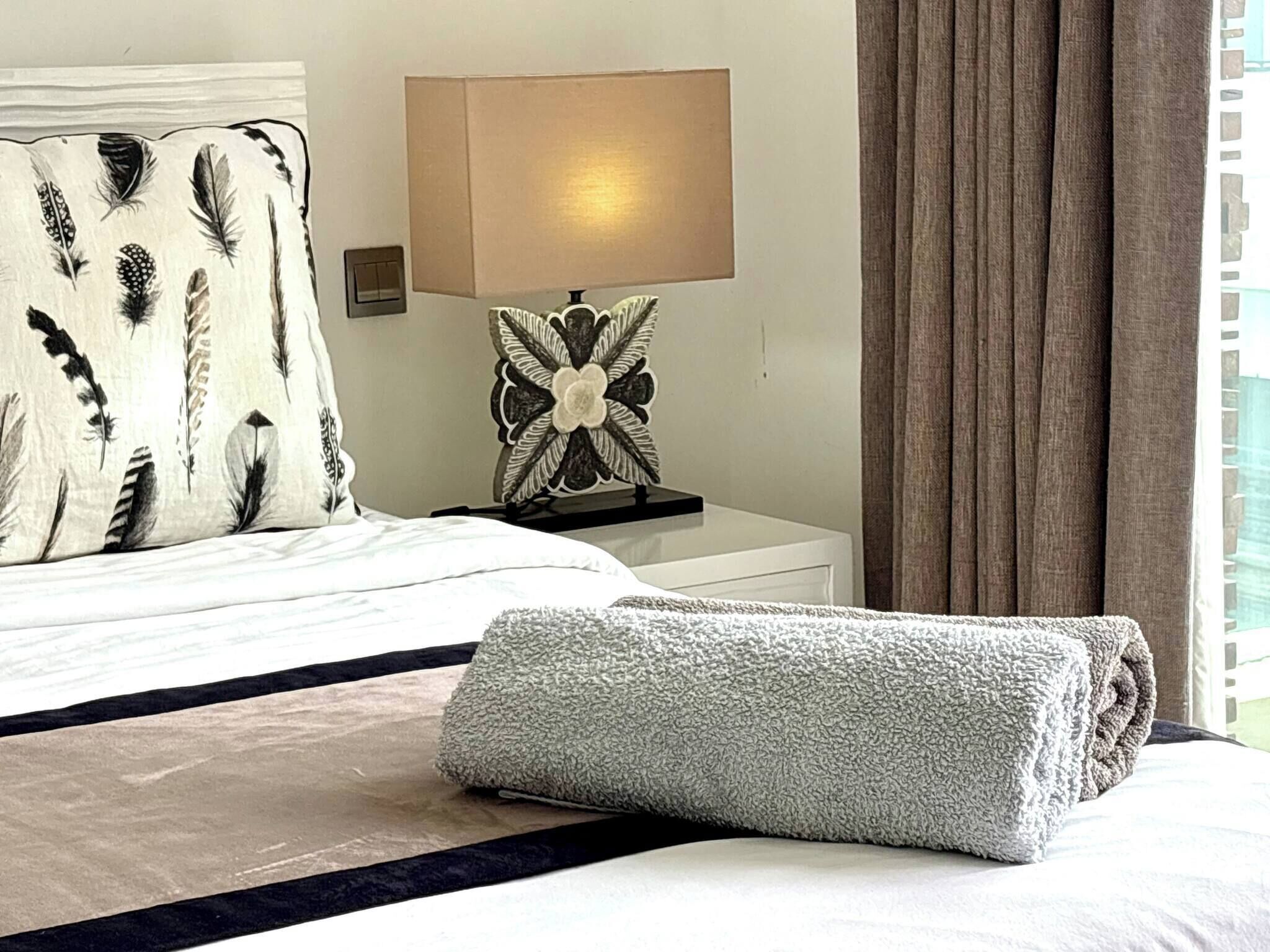 A neatly made bed with a white and black striped bedspread, a grey folded blanket, a decorative lamp with a floral design, and a window with curtains.