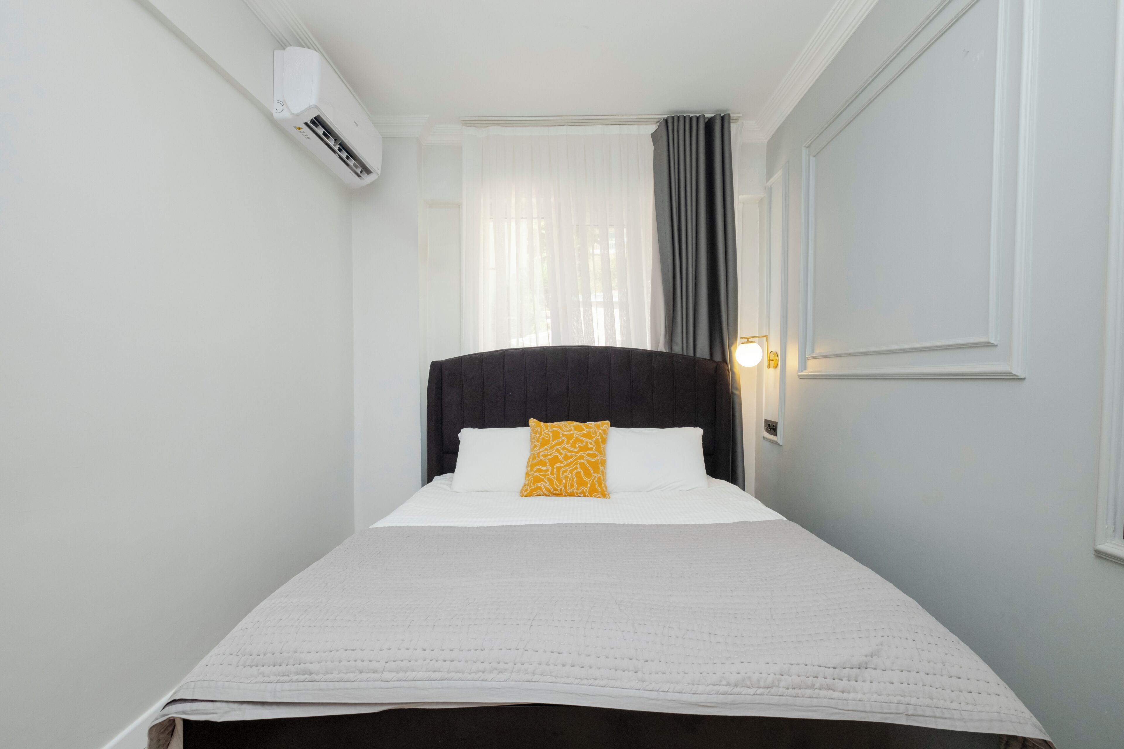 Une chambre d’hôtel étroite, avec un lit simple, du linge de lit blanc, une tête de lit sombre, une fenêtre avec des rideaux légers et un climatiseur fixé au mur.
