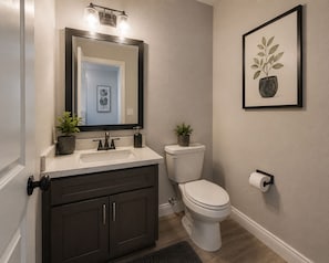 Ein Badezimmer mit einem dunklen Holz-Waschtisch, einer weißen Toilette, einem Waschbecken mit Spiegel darüber und einer gerahmten Pflanze an der Wand.