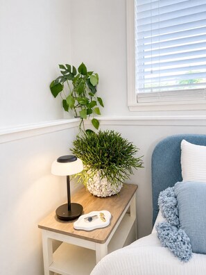 Un dormitorio con una mesita de noche, una lámpara, una planta en maceta, una cama con cabecera azul y una ventana con persianas.