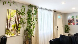 A living room with a large painting, a potted plant, a window with curtains, and a door. - Cozy Seattle Suite Near SeaTac Airport & Downtown Seattle (Burien)